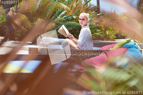 Image of Candid shot of lady reading book and relaxing in lush tropical garden.