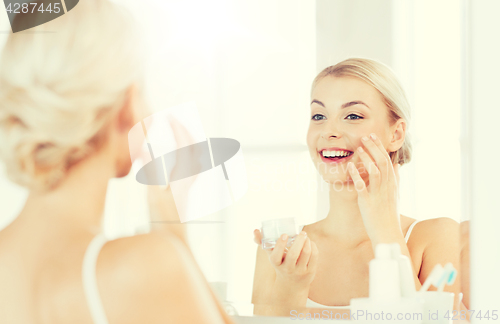 Image of happy woman applying cream to face at bathroom