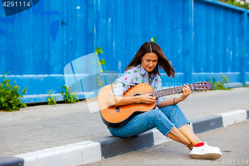 Image of woman in jeans sits on a road curb and plays the guitar