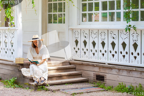 Image of woman in a white dress and a straw hat reading a book on the por