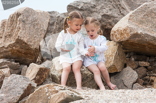 Image of Children on the sea beach. Twins sitting against stones and sea water.