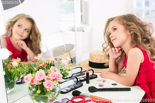 Image of A little girl with cosmetics. She is in mother\'s bedroom, sitting near the mirror.