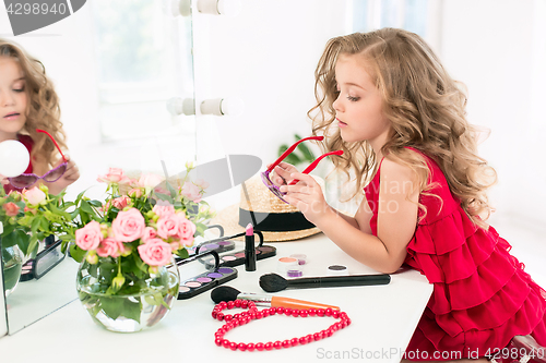 Image of A little girl with cosmetics. She is in mother\'s bedroom, sitting near the mirror.