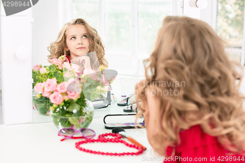 Image of A little girl with cosmetics. She is in mother\'s bedroom, sitting near the mirror.