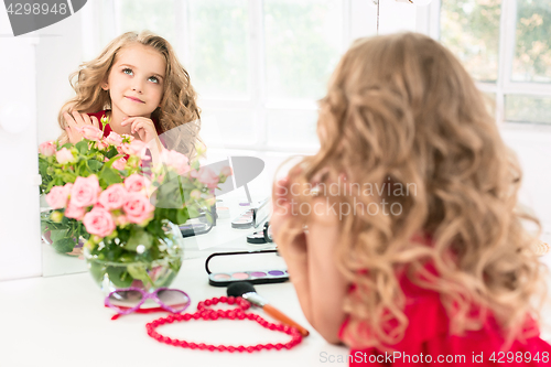 Image of A little girl with cosmetics. She is in mother\'s bedroom, sitting near the mirror.