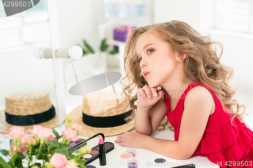 Image of A little girl with cosmetics. She is in mother\'s bedroom, sitting near the mirror.