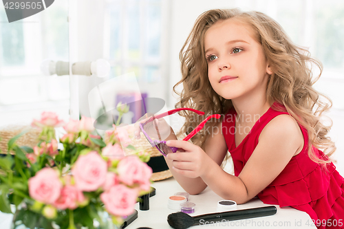 Image of A little girl with cosmetics. She is in mother\'s bedroom, sitting near the mirror.