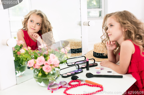 Image of A little girl with cosmetics. She is in mother\'s bedroom, sitting near the mirror.