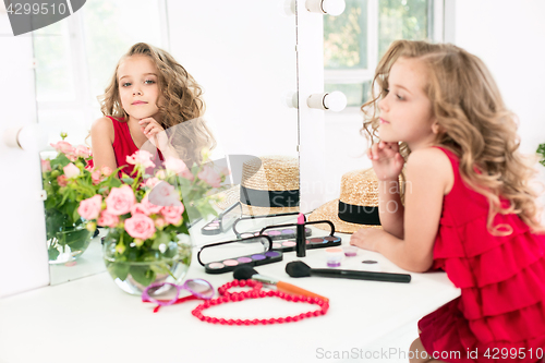 Image of A little girl with cosmetics. She is in mother\'s bedroom, sitting near the mirror.