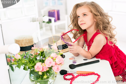 Image of A little girl with cosmetics. She is in mother\'s bedroom, sitting near the mirror.