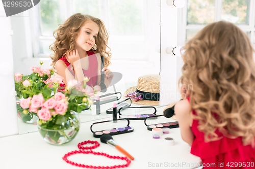 Image of A little girl with cosmetics. She is in mother\'s bedroom, sitting near the mirror.