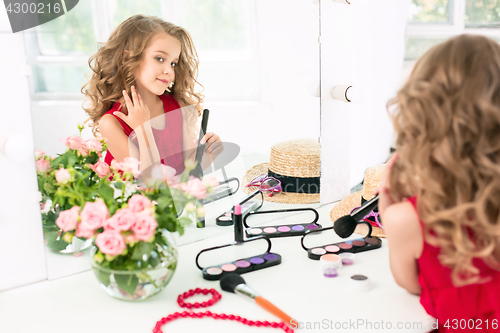 Image of A little girl with cosmetics. She is in mother\'s bedroom, sitting near the mirror.