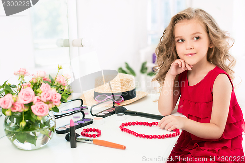 Image of A little girl with cosmetics. She is in mother\'s bedroom, sitting near the mirror.