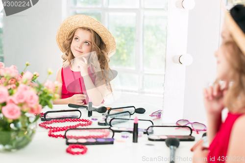 Image of A little girl with cosmetics. She is in mother\'s bedroom, sitting near the mirror.