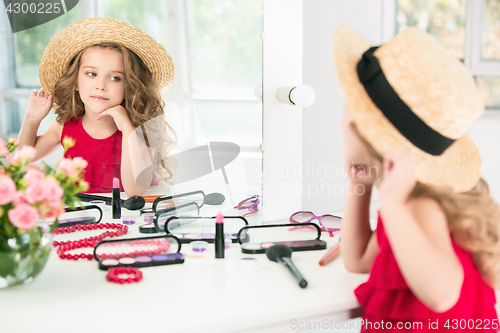 Image of A little girl with cosmetics. She is in mother\'s bedroom, sitting near the mirror.