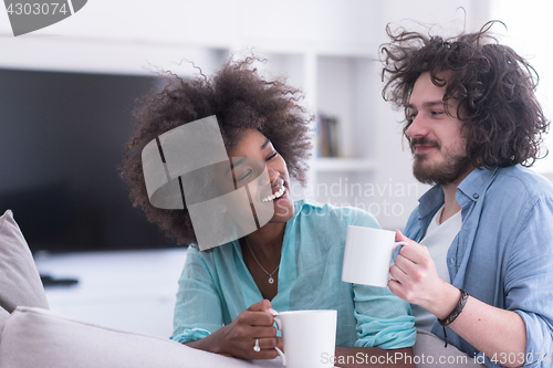 Image of multiethnic couple sitting on sofa at home drinking coffe