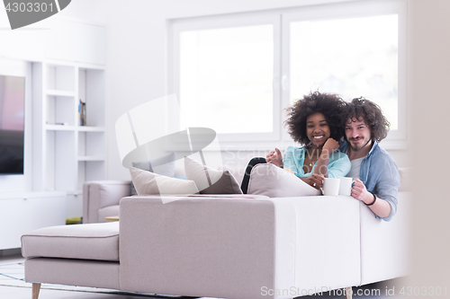 Image of multiethnic couple sitting on sofa at home drinking coffe