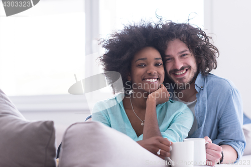 Image of multiethnic couple sitting on sofa at home drinking coffe