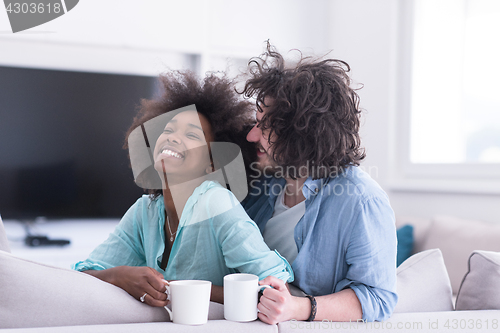 Image of multiethnic couple sitting on sofa at home drinking coffe
