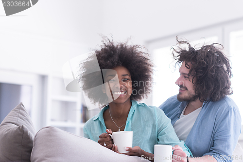 Image of multiethnic couple sitting on sofa at home drinking coffe