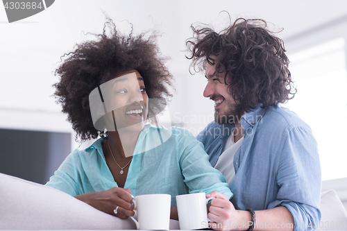 Image of multiethnic couple sitting on sofa at home drinking coffe