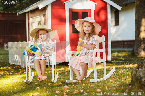Image of Cute little blond girls in hats sitting on the field with soft toys in summer.