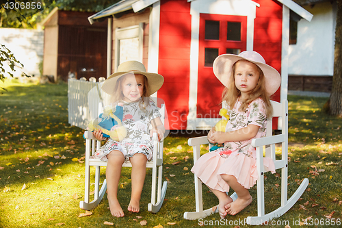 Image of Cute little blond girls in hats sitting on the field with soft toys in summer.