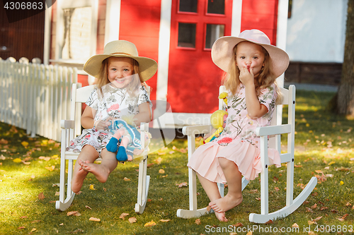 Image of Cute little blond girls in hats sitting on the field with soft toys in summer.