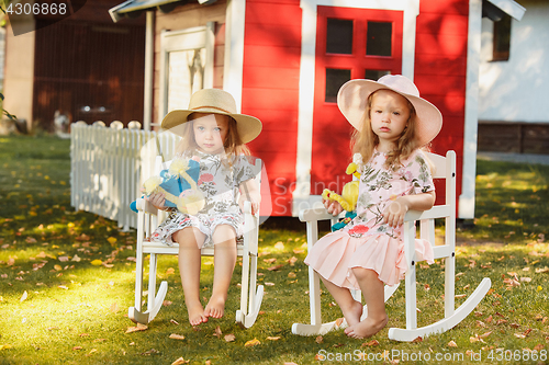Image of Cute little blond girls in hats sitting on the field with soft toys in summer.