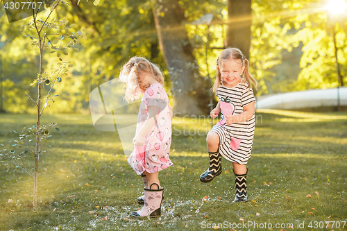 Image of The cute little blond girls in rubber boots playing with water splashes on the field in summer