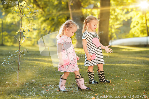 Image of The cute little blond girls in rubber boots playing with water splashes on the field in summer