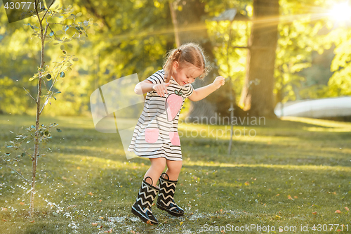 Image of The cute little blond girl in rubber boots playing with water splashes on the field in summer