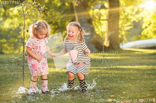 Image of The cute little blond girls in rubber boots playing with water splashes on the field in summer