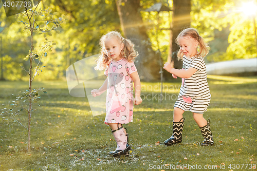 Image of The cute little blond girls in rubber boots playing with water splashes on the field in summer