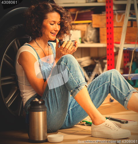 Image of woman in blue overalls sitting near the wheel of a car and drink