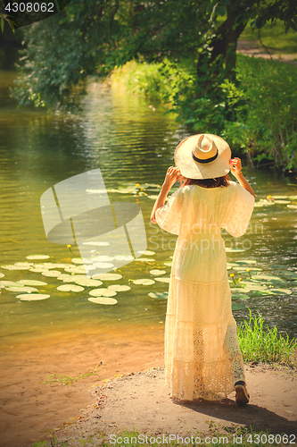 Image of girl in a white dress and hat on the shore of a pond with water-