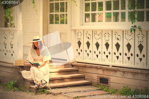 Image of woman in a white dress and a straw hat reading a book on the por