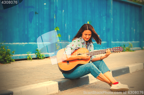 Image of woman in jeans sits on a road curb and plays the guitar