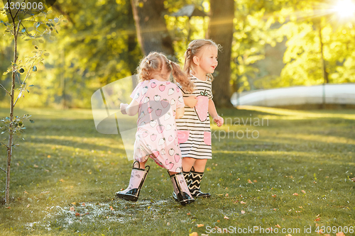 Image of The cute little blond girls in rubber boots playing with water splashes on the field in summer