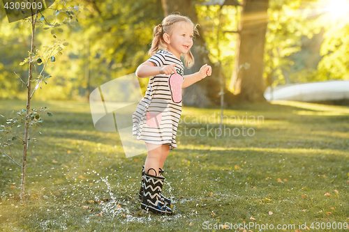 Image of The cute little blond girl in rubber boots playing with water splashes on the field in summer