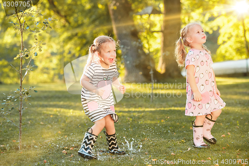 Image of The cute little blond girls in rubber boots playing with water splashes on the field in summer