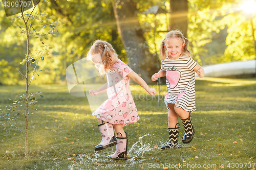 Image of The cute little blond girls in rubber boots playing with water splashes on the field in summer