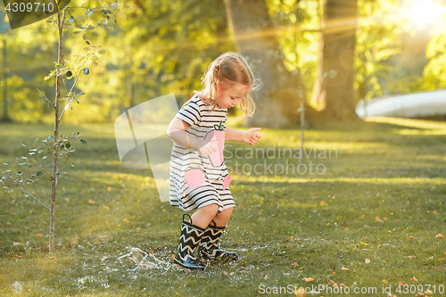 Image of The cute little blond girl in rubber boots playing with water splashes on the field in summer