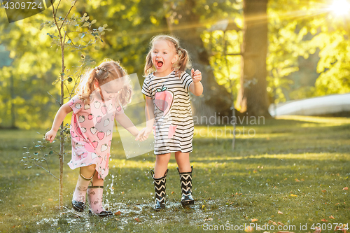 Image of The cute little blond girls in rubber boots playing with water splashes on the field in summer