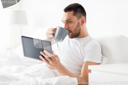 Image of man with tablet pc drinking coffee in bed at home