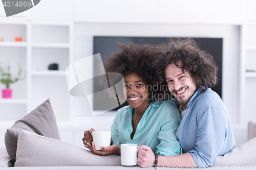 Image of multiethnic couple sitting on sofa at home drinking coffe