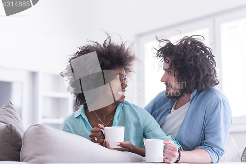 Image of multiethnic couple sitting on sofa at home drinking coffe