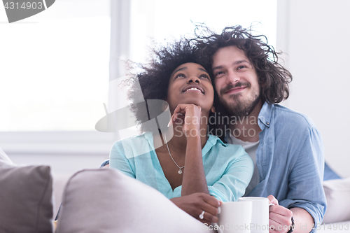 Image of multiethnic couple sitting on sofa at home drinking coffe