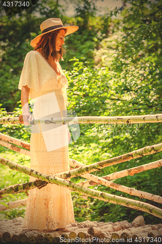 Image of woman in a white dress and a summer hat on a bridge from a birch