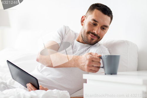 Image of man with tablet pc drinking coffee in bed at home